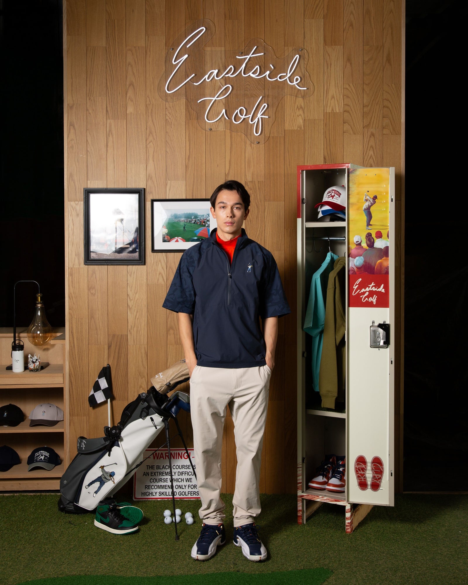 Eastside Golf campaign model in a navy pullover and cream trousers beneath the brand’s neon sign in a golf locker-room set